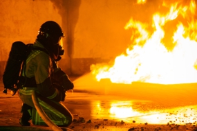 links kniet ein Feuerwehrmann in Schutzkleidung und löscht einen Brandherd am Boden