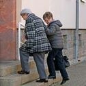 Im Bild eine ältere Dame mit einer Betreuerin, die ihr hilft beim Treppensteigen
